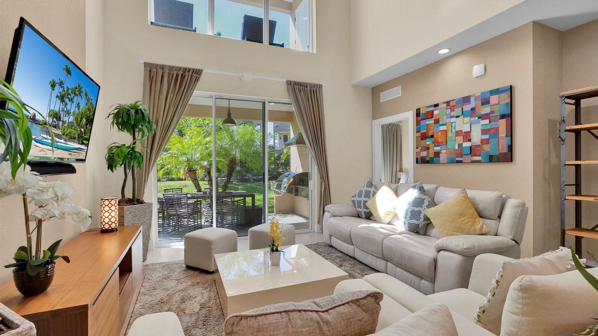 A spacious living room with a large sofa, a coffee table, and a TV on the left. There is a sliding glass door leading to a balcony with outdoor furniture.