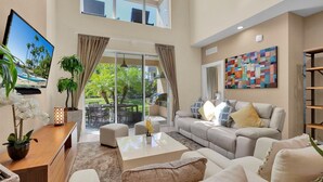 A spacious living room with a large sofa, a coffee table, and a TV on the left. There is a sliding glass door leading to a balcony with outdoor furniture. - Magic Village Resort 4 Bedroom townhome with Outdoor Kitchen 4 Townhouse (Kissimmee)