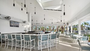 A modern bar area with white bar stools and a long counter, large windows with a view of palm trees, and ceiling fans. - Windsor Cay Resort 5 Bedroom Townhome with Pool 5 Townhouse (Clermont)