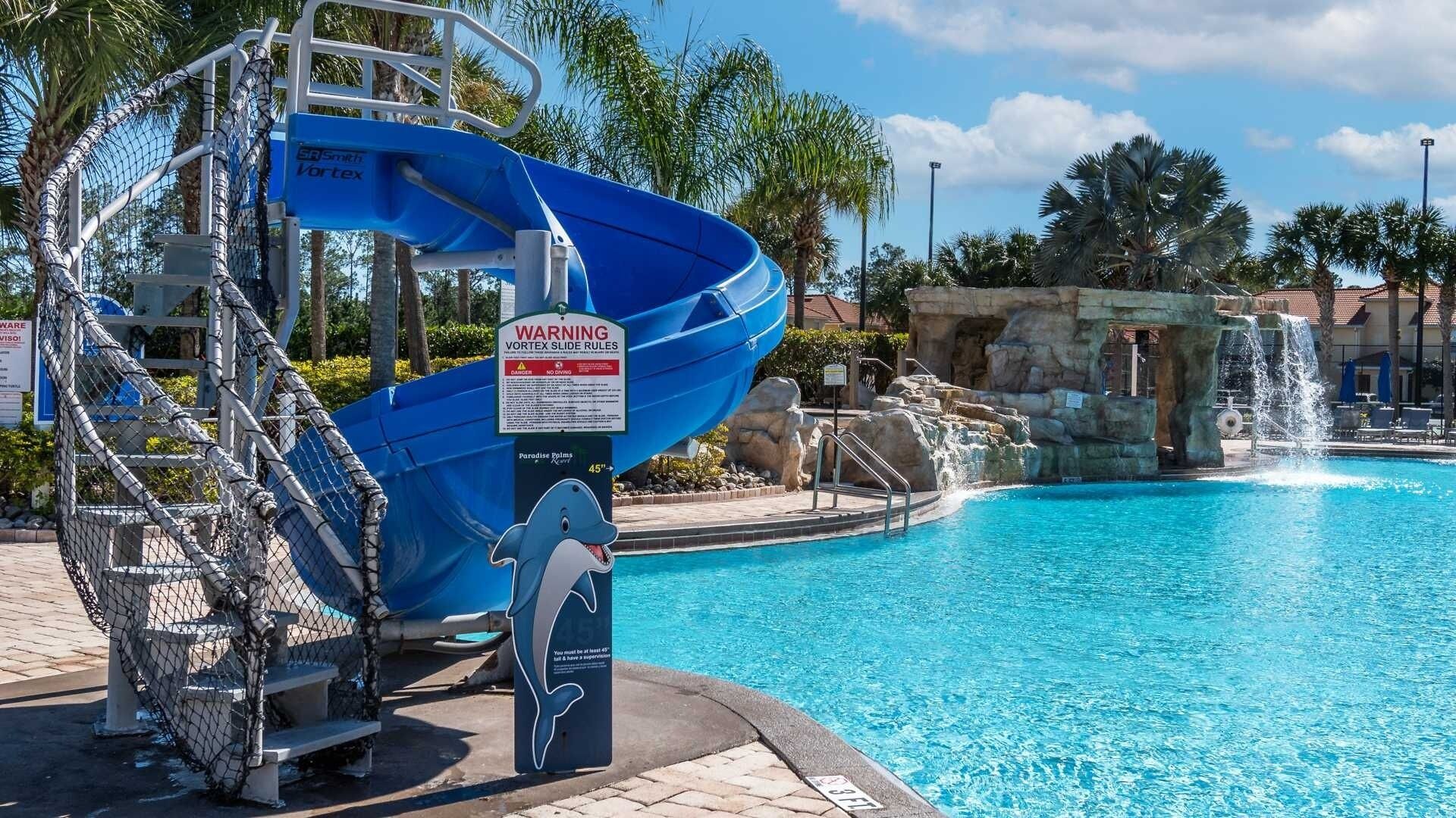 Une toboggan aquatique bleu au bord d’une piscine, avec un panneau d’avertissement, une sculpture de dauphin et une cascade décorative.