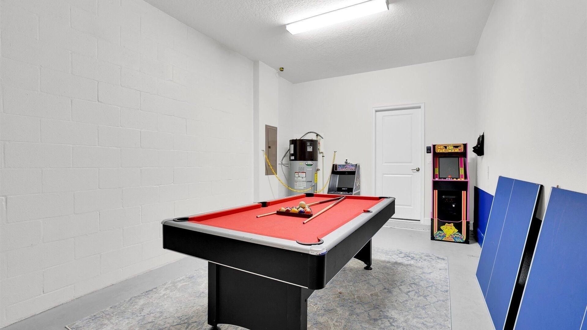 A pool table with balls and cues, a water heater, and a pinball machine in a room with white brick walls and a door.