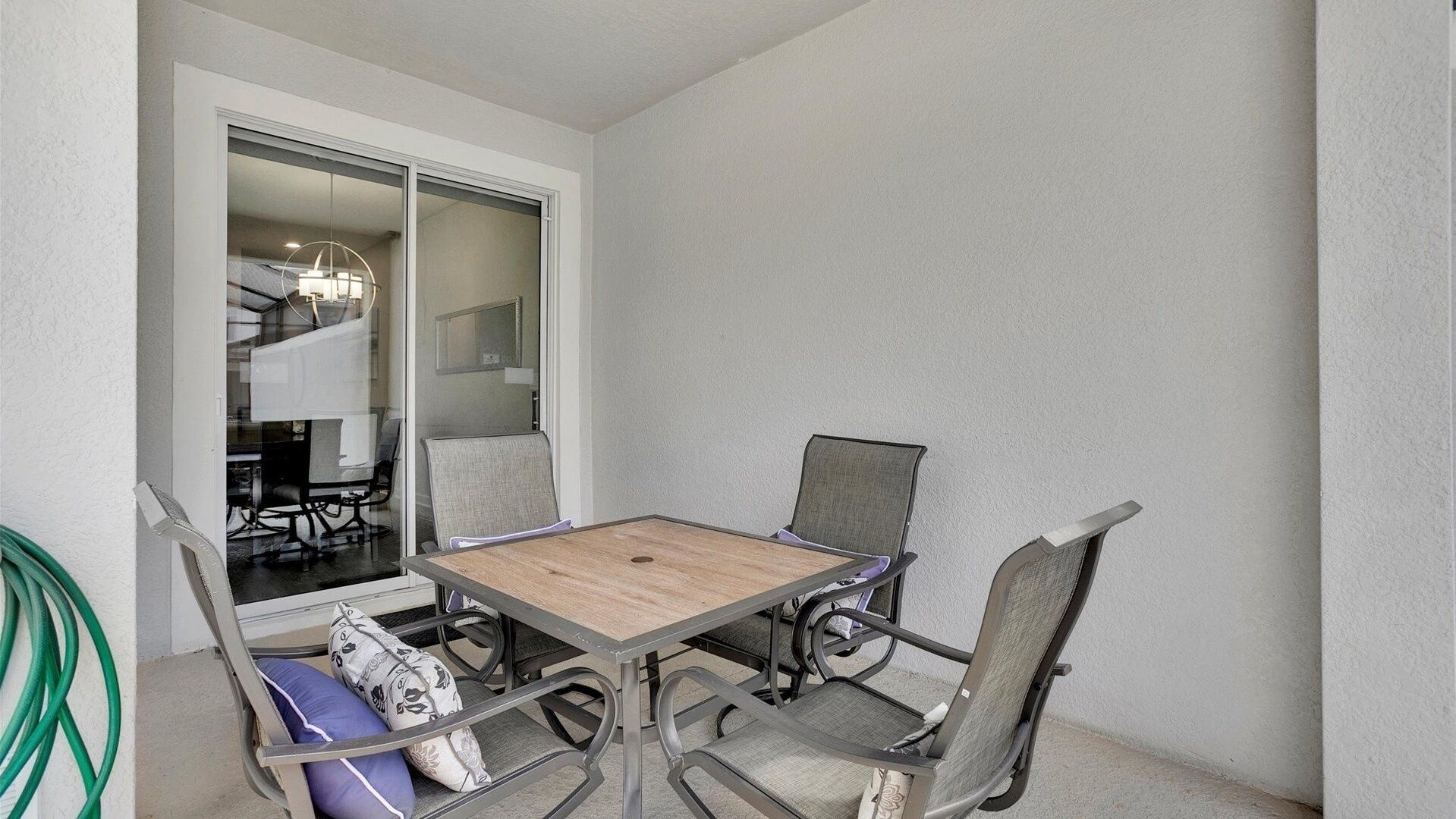 Een klein terras met een tafel en vier stoelen, een schuifdeur naar een eethoek, en een groene tuinslang aan de linkerkant.