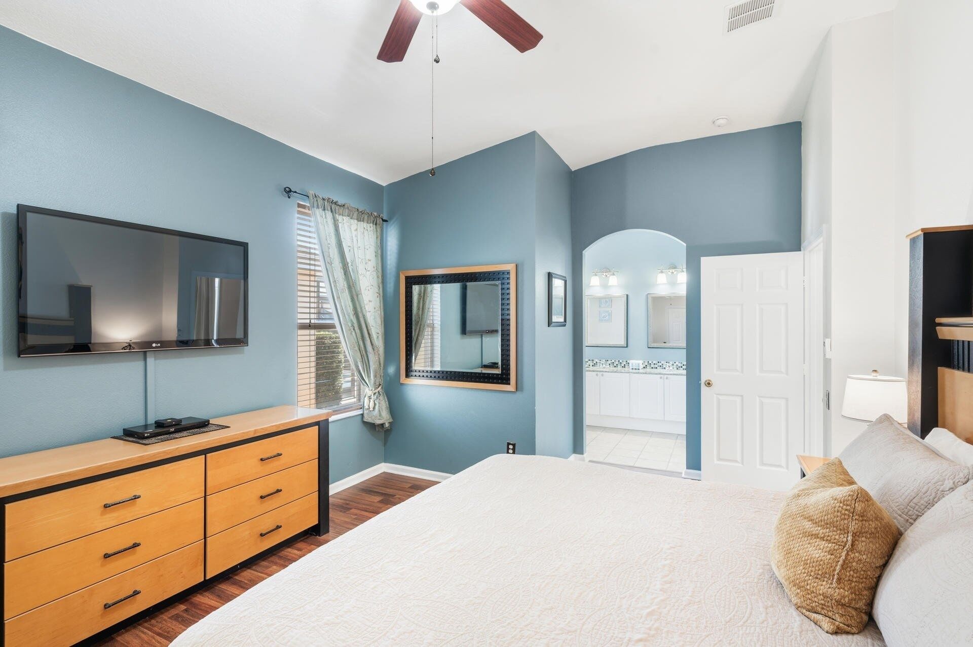 Une chambre à coucher comprenant un lit, une commode, un téléviseur, un ventilateur de plafond, et une salle de bain visible à travers une arche.