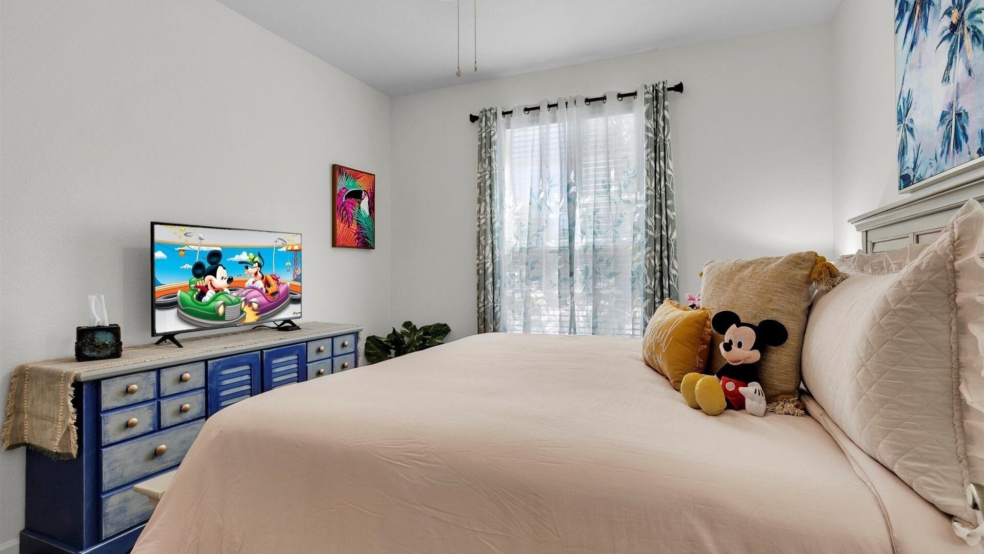 A bedroom with a bed, a TV, a Mickey Mouse plush toy, and a painting on the wall.