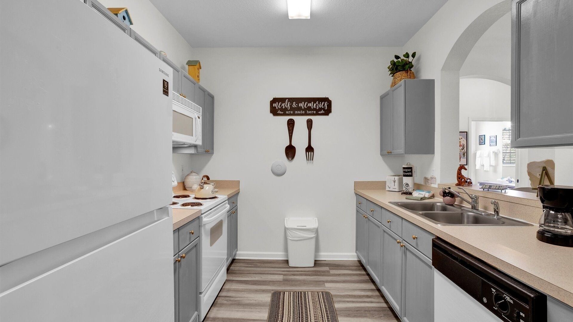 A modern kitchen with grey cabinets, a white countertop, and a wooden floor. A sign on the wall reads "meals & memories are made here."