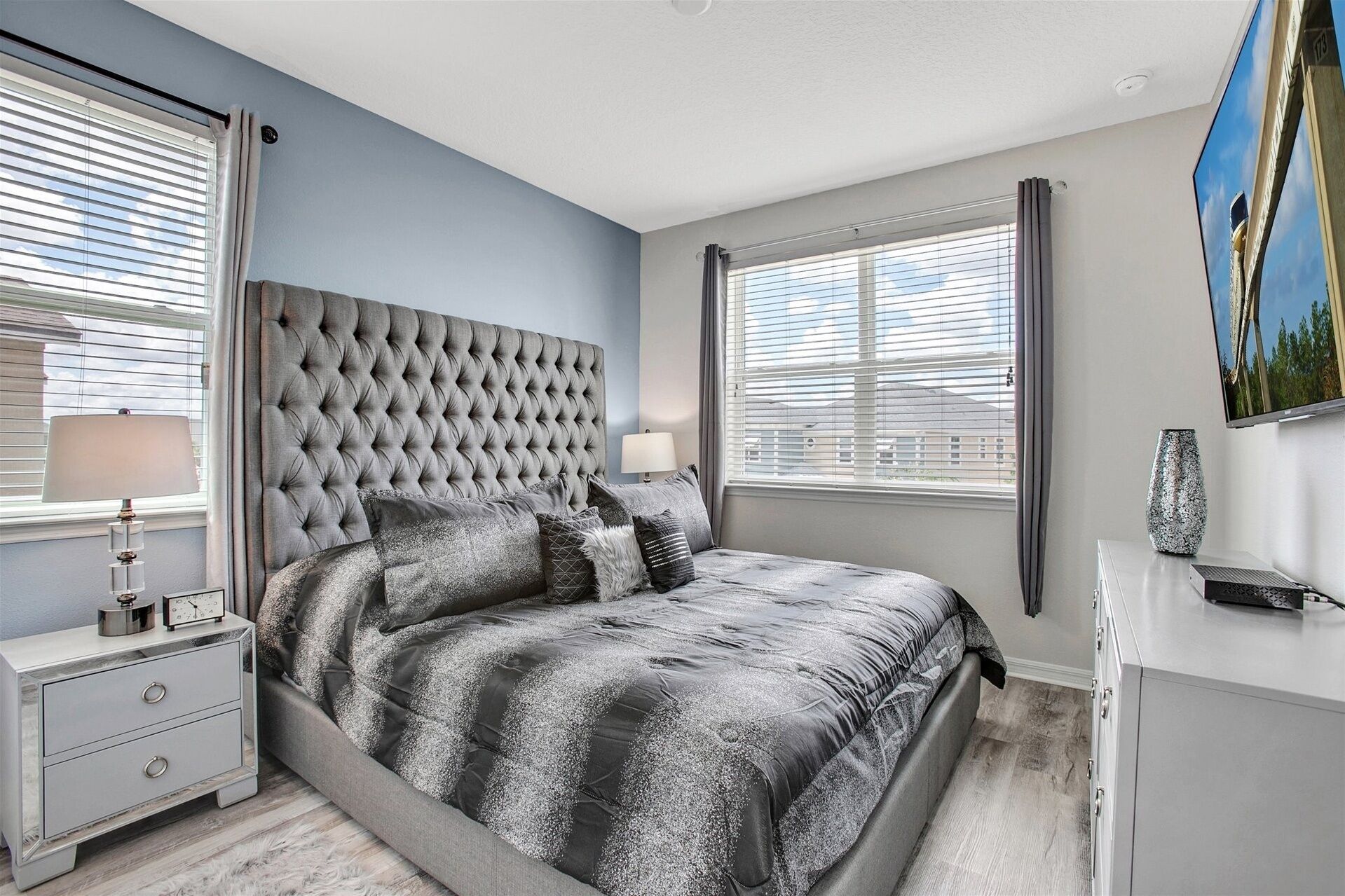 A bedroom with a tufted headboard, a bed with a patterned duvet, bedside lamps, a nightstand, a dresser, and a painting on the wall.