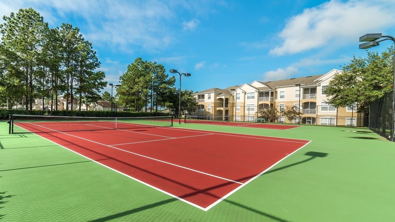 Cancha de tenis con red, rodeada de árboles y un edificio con balcones.