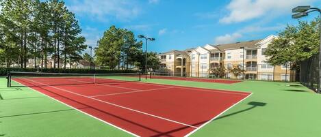 Cancha de tenis con red, rodeada de árboles y un edificio con balcones.