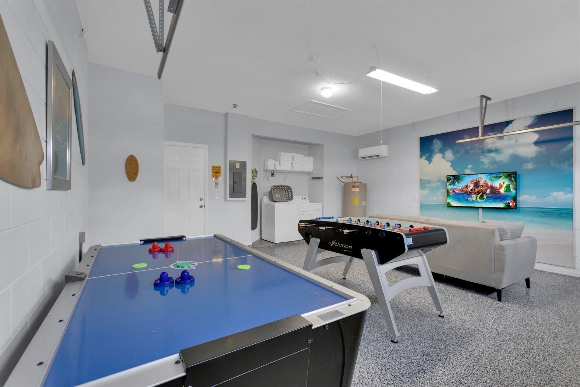 A room with two foosball tables, a sofa, a television, and a painting of a beach scene.