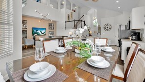 Une salle à manger spacieuse, avec une grande table en bois dressée pour six personnes, entourée de chaises blanches. Un escalier avec une rampe noire mène à l’étage supérieur.