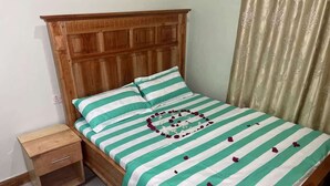 A single bed with a wooden headboard, a small bedside table, and a green and white striped bedspread with a heart-shaped decoration made of rose petals.