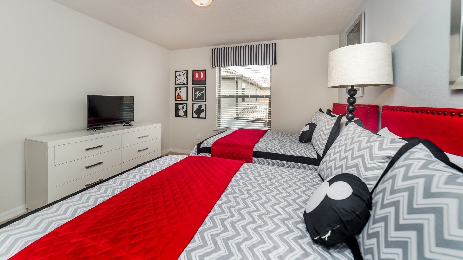 A bedroom with two beds, a TV on a white dresser, and a window with blinds.