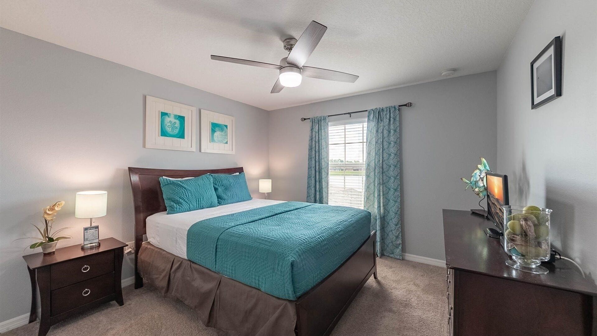 Une chambre à coucher comprenant un lit, une table de chevet, une commode, un ventilateur de plafond et une fenêtre avec des rideaux.