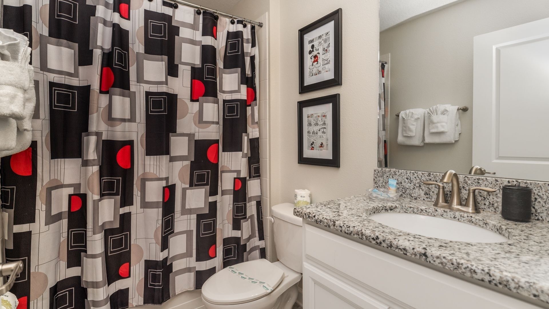 Een badkamer met een douchegordijn met patroon, een toilet, een wastafel met een granieten aanrechtblad en ingelijste foto's aan de muur.