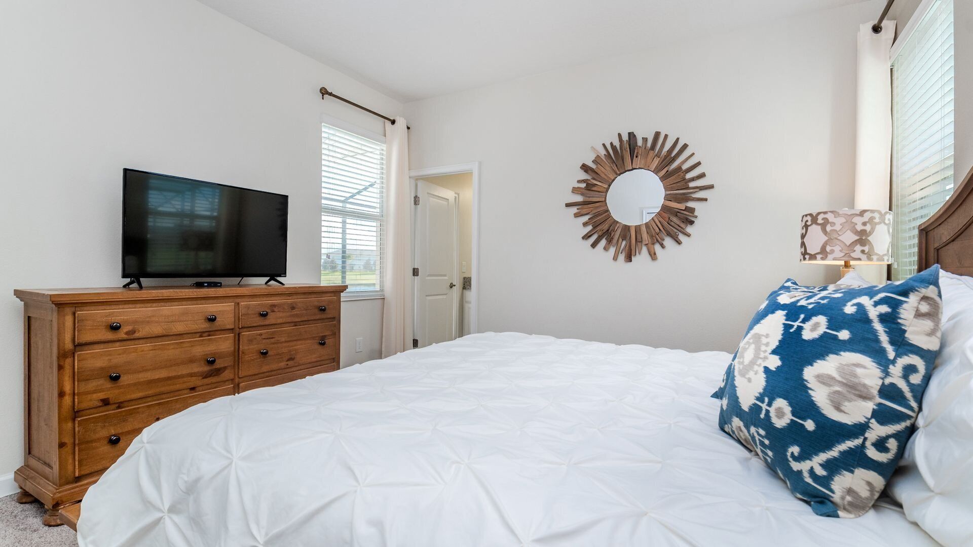 A bedroom with a bed, a wooden dresser, a television, a mirror, and a window with blinds.