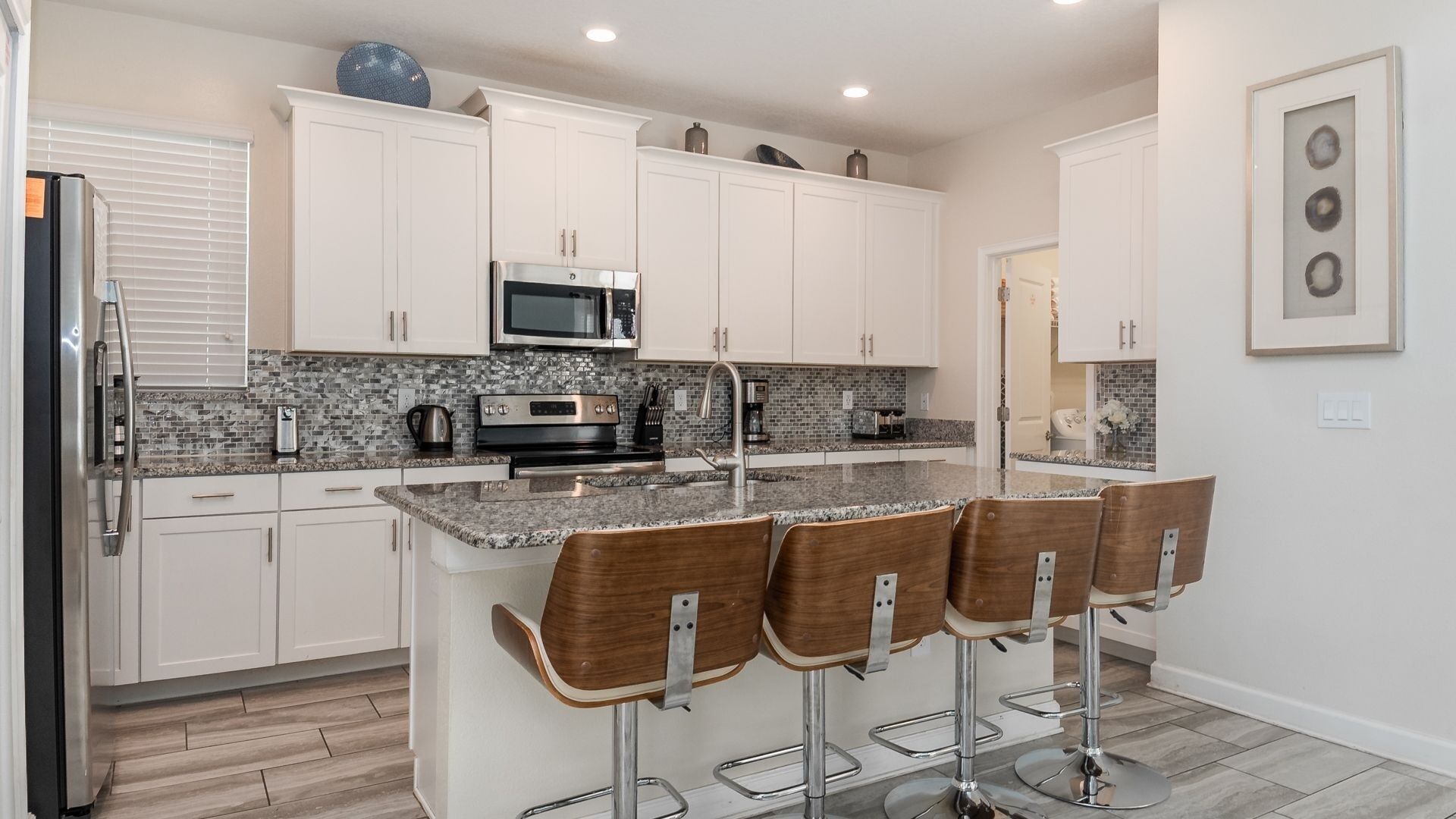A modern kitchen with white cabinets, a granite countertop, a stainless steel refrigerator, a microwave, and a coffee maker. There are four wooden bar stools with metal legs.