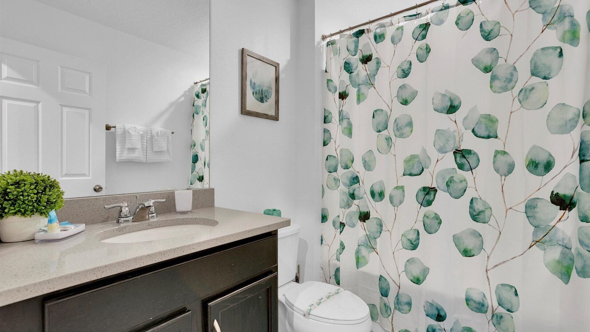 A bathroom with a white sink, a mirror, a framed picture on the wall, and a shower curtain with a leaf pattern.