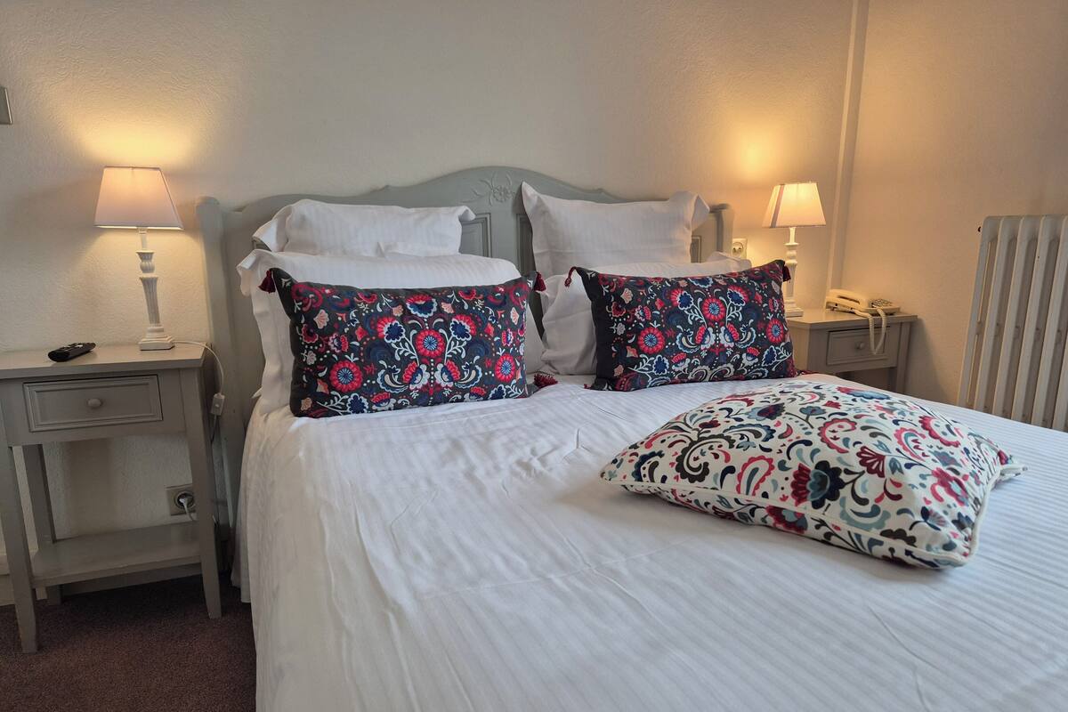 A neatly made bed with white linens and decorative pillows, a bedside table with a lamp, and a bedside phone.