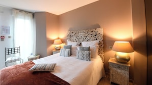 A neatly made bed with a patterned headboard, two bedside lamps, and a chair with a cushion. - LA VILLA JULIA (Agon-Coutainville)