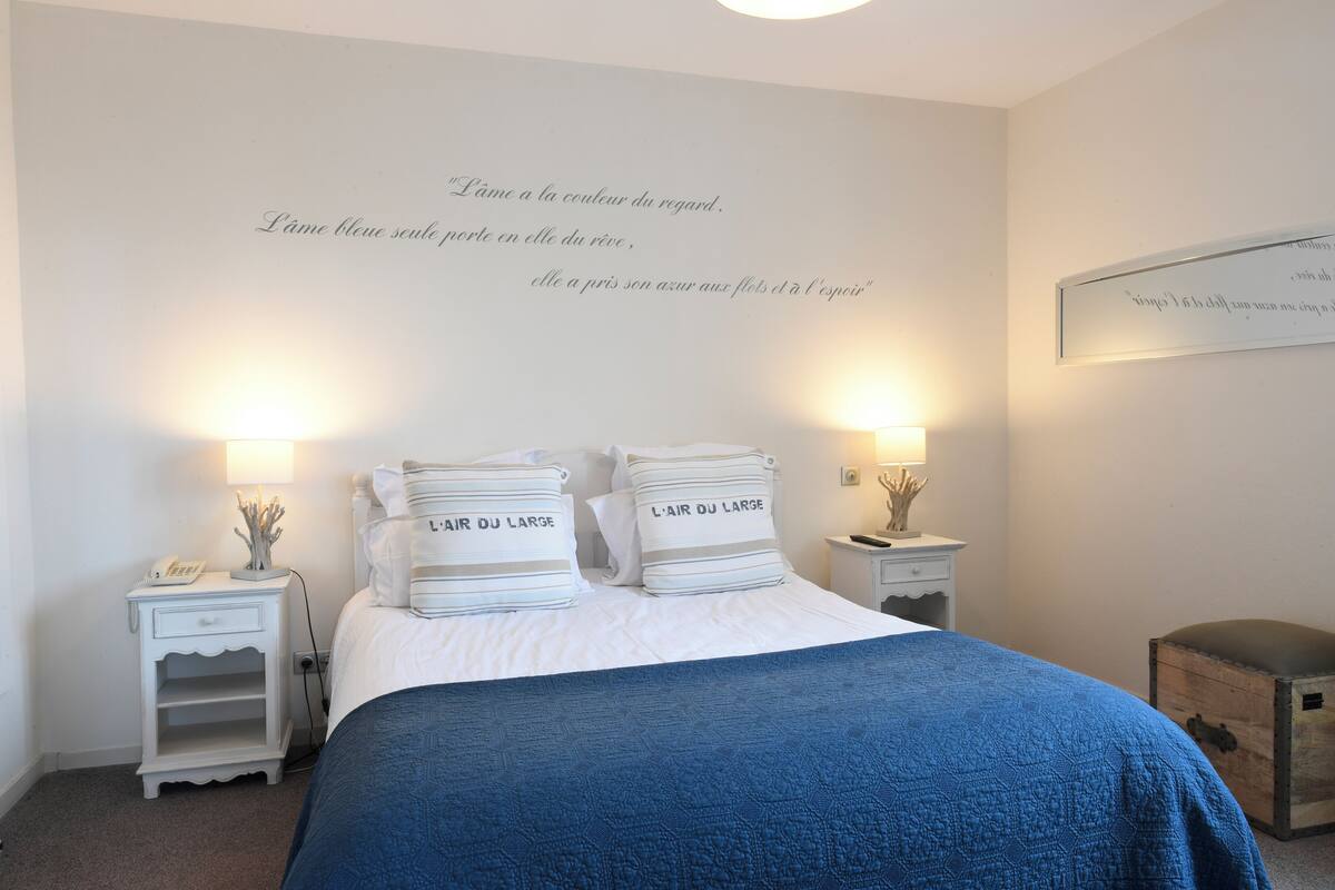 A bedroom with a bed, two bedside tables, a nightstand, a blue blanket, and a wall with French text.