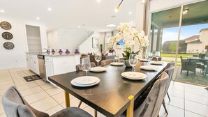 A modern dining area with a dark wooden table set for six, surrounded by grey upholstered chairs. A large window offers a view of the outdoors. - Solara Resort 6 Bedroom Private Pool+Spa Home 6 Home (Kissimmee)