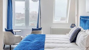 A hotel room with a bed, a chair, a small table, a chandelier, and a view of the city through the windows. - Beachfront Luxury Home with Hot Tub, 9 beds, Cinema room, Sleeps 2 to 16 (Newquay)