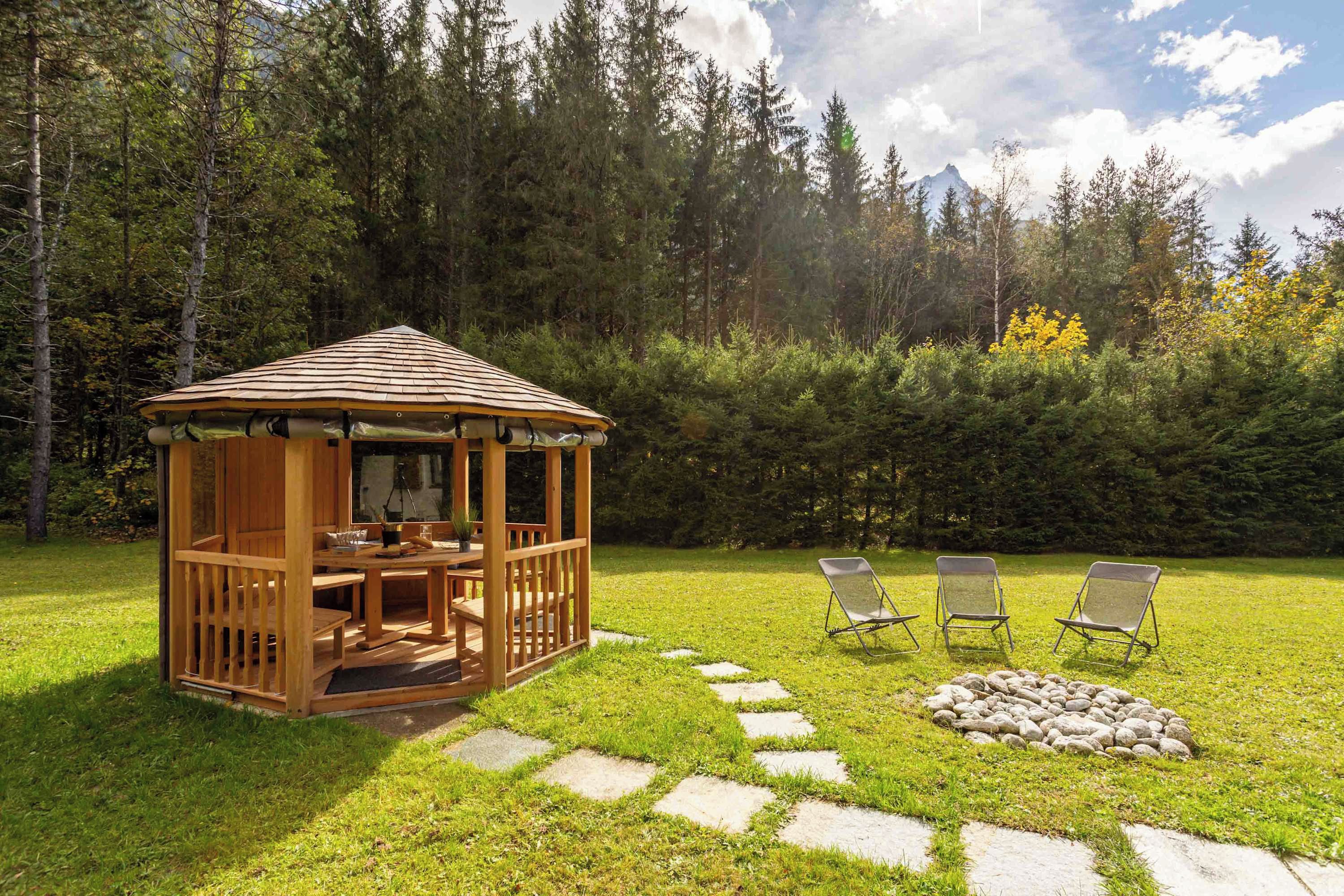 A wooden gazebo with a table and chairs in a grassy area, surrounded by trees and a stone path.