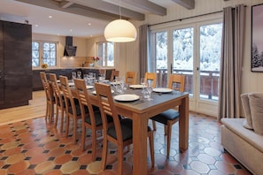 A dining area with a wooden table set for six, surrounded by chairs, in a room with a hexagonal tile floor and large windows. - Chalet le Petit Tricouni, ski, Chamonix All Year (Chamonix)