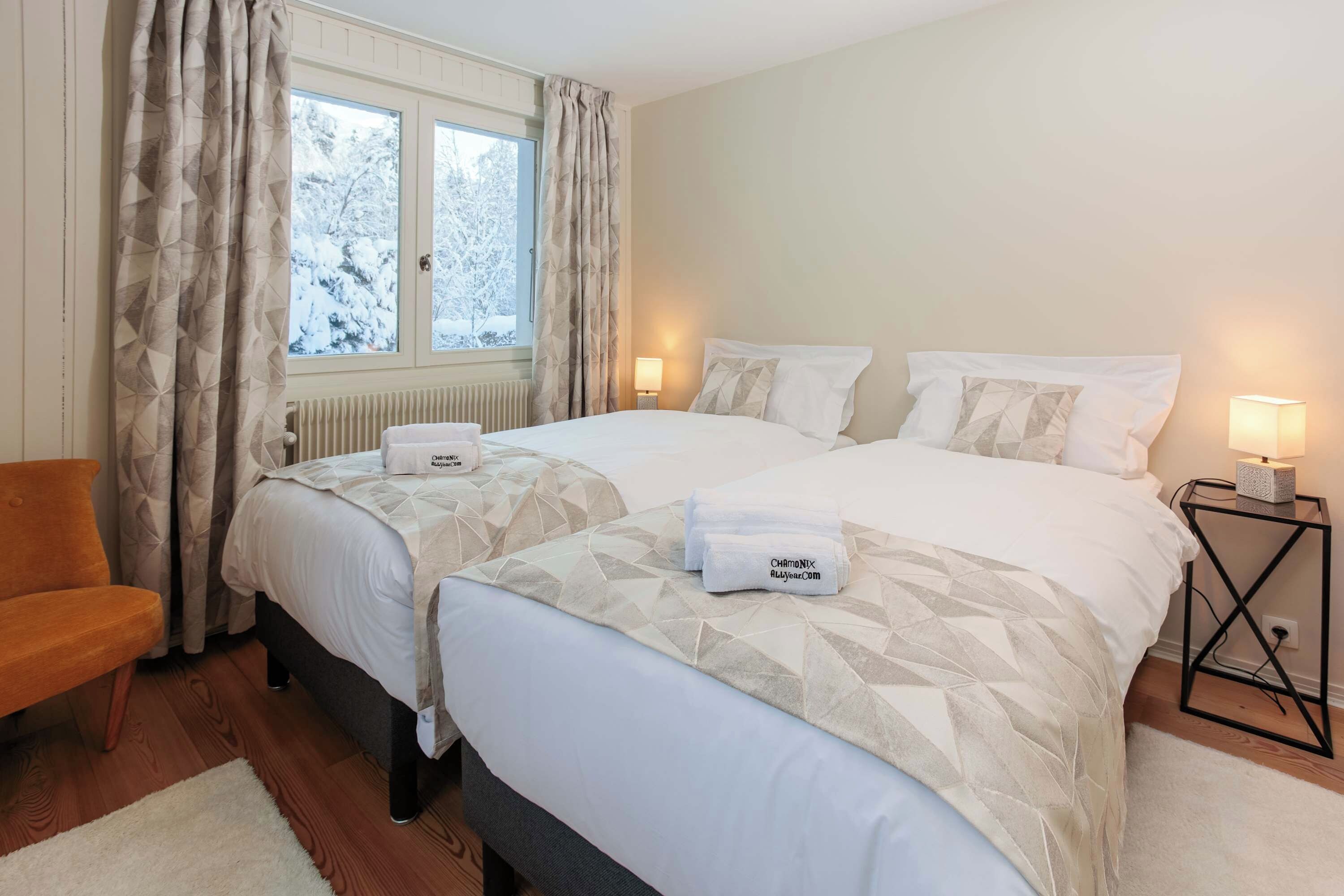 A hotel room with two beds, a chair, a bedside table with a lamp, and a view of snow-covered trees through the window.