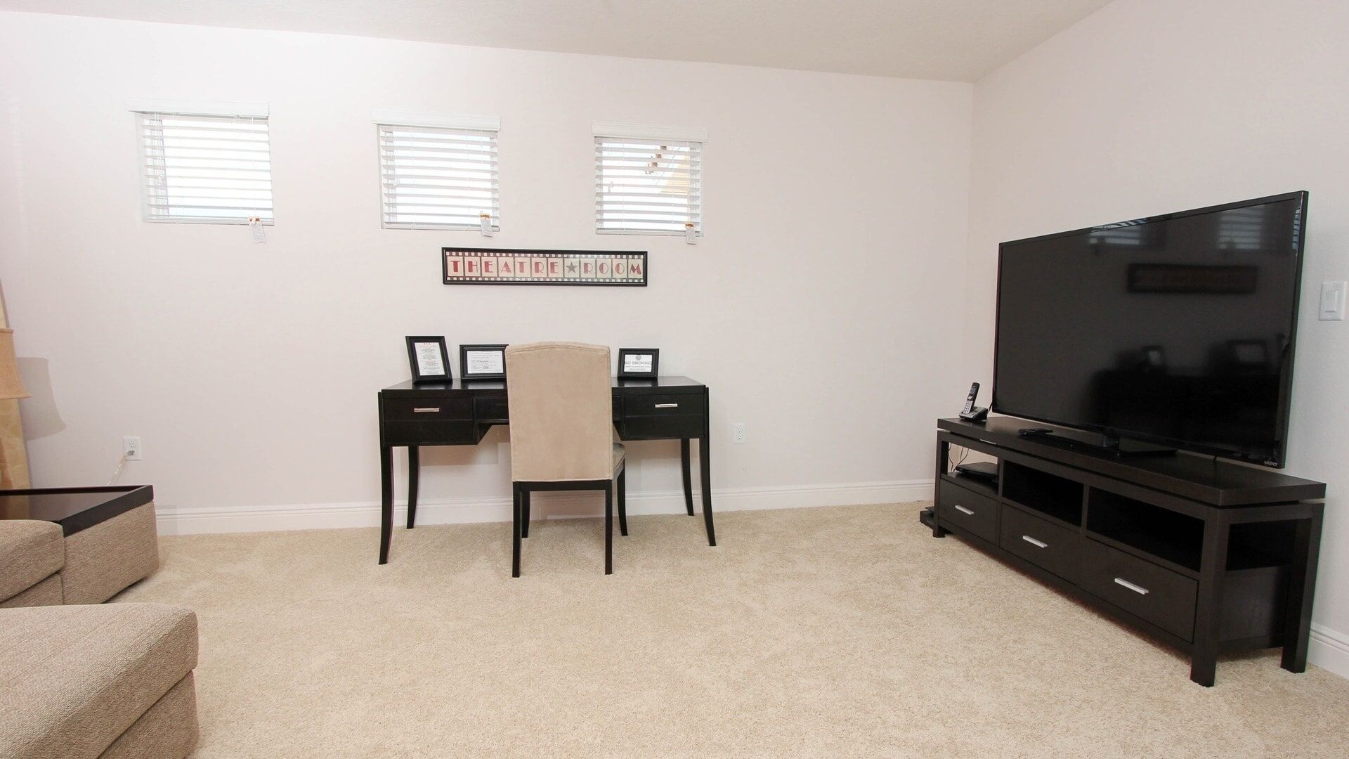 A living room with a black entertainment center, a large flat-screen TV, a beige sofa, and a desk with two chairs. There are three windows with blinds and a framed sign on the wall.