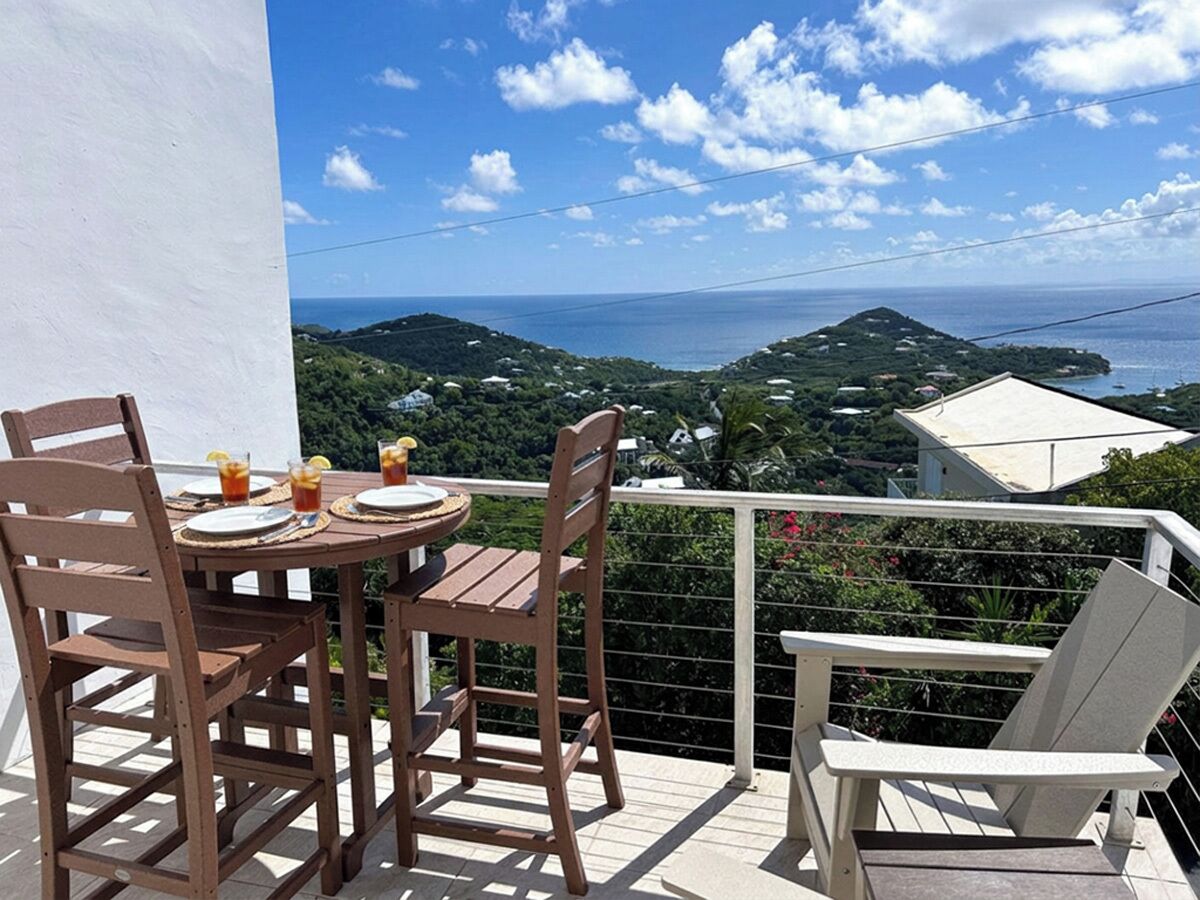 A balcony with a table and chairs, overlooking a coastal landscape with hills and a view of the sea.