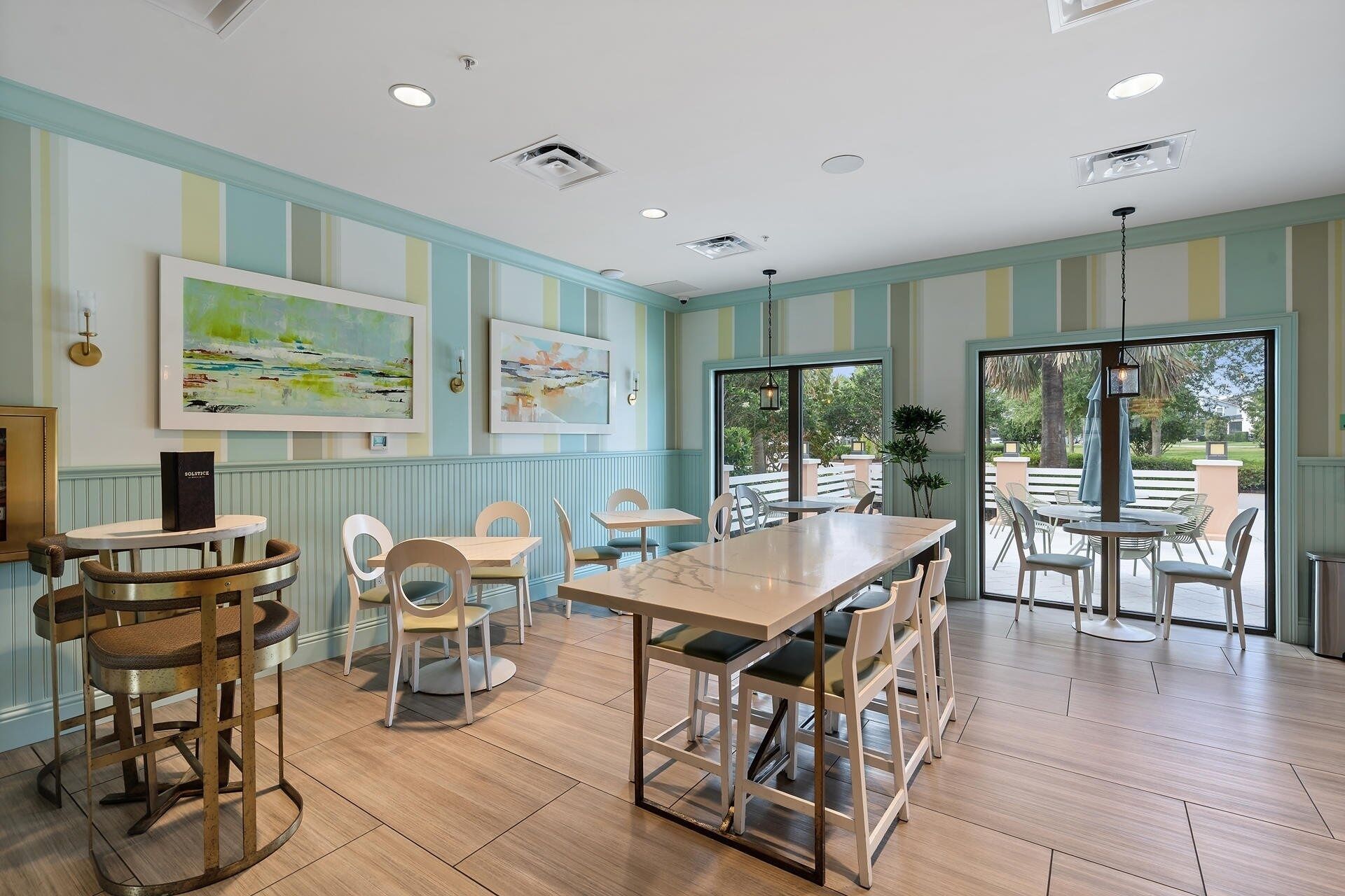 A dining area with wooden tables and chairs, artwork on the walls, and a view of the outdoors through large windows.