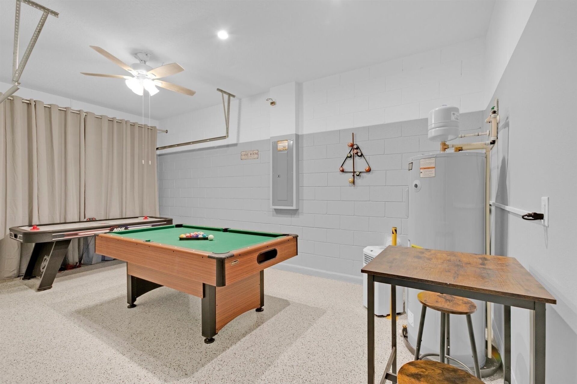 A pool table with a ceiling fan, a billiards rack, and a table with stools in a room with white walls and a tiled floor.