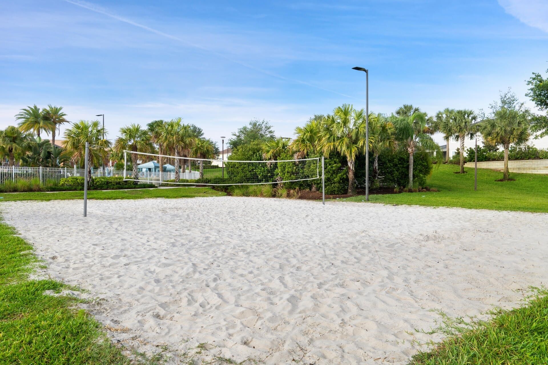 A sandy beach volleyball court with a net, surrounded by palm trees and a fence.