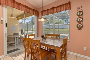 Un espace repas avec une table en marbre et des chaises en bois, un ventilateur de plafond et une vue dégagée grâce à de grandes fenêtres équipées de stores.
