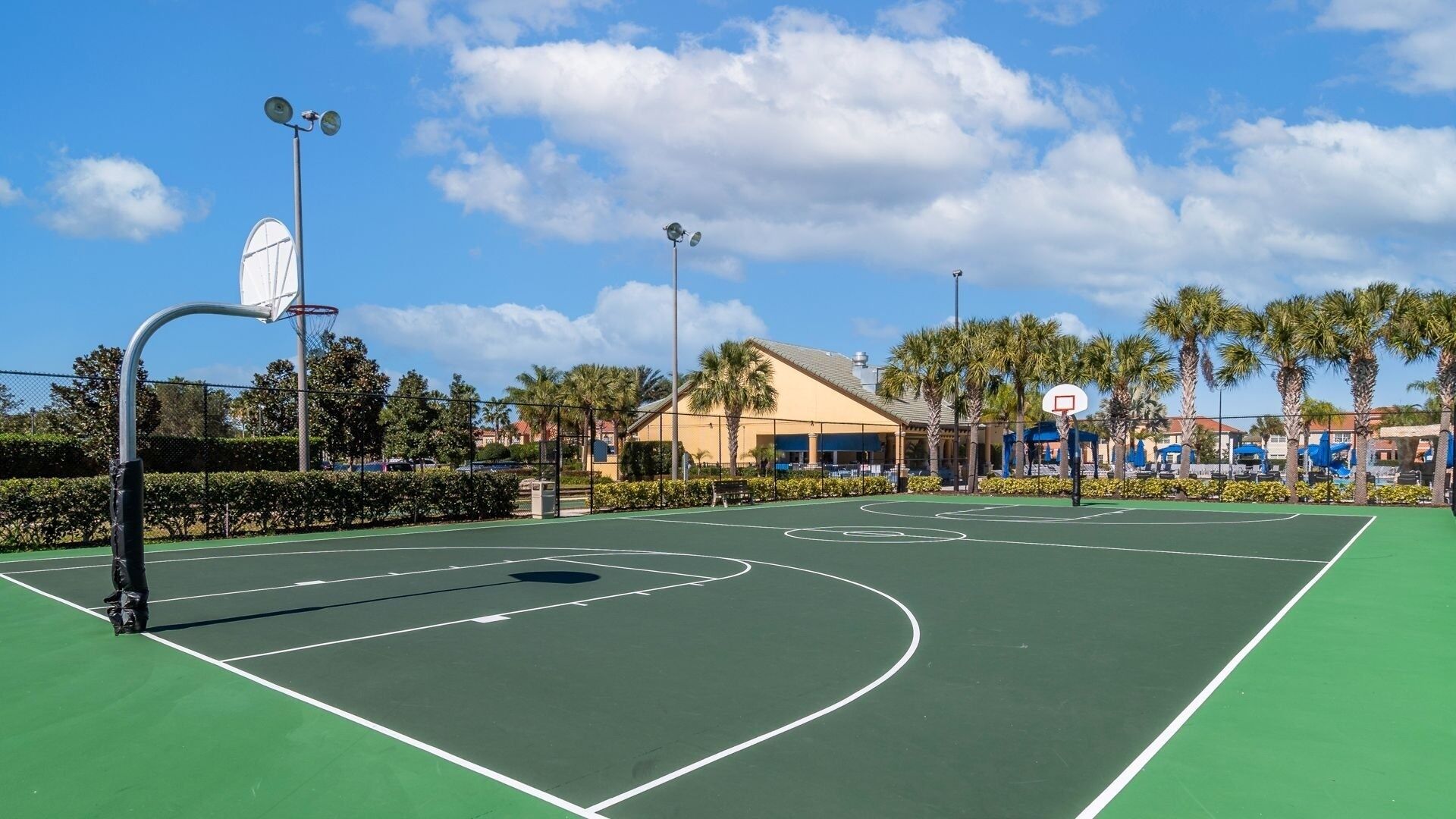 Un terrain de basket en plein air, doté d’une surface verte, entouré de palmiers et bordé d’un bâtiment au toit rouge.