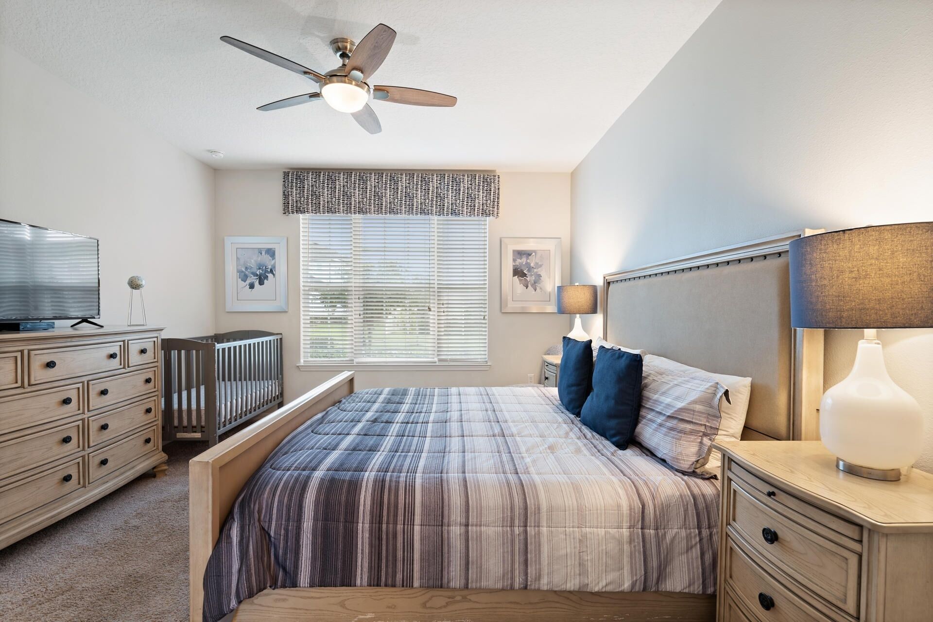 A bedroom with a bed, bedside tables, a ceiling fan, a TV, and a window with blinds.