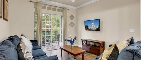 A living room with a ceiling fan, a large window with curtains, a wooden coffee table, and a television on the wall.