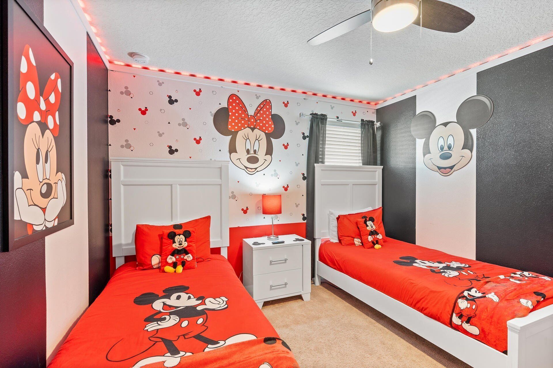 A room with two beds, each with red bedding featuring Mickey Mouse designs. There are wall decals of Mickey and Minnie Mouse, a ceiling fan, and a nightstand with a lamp.