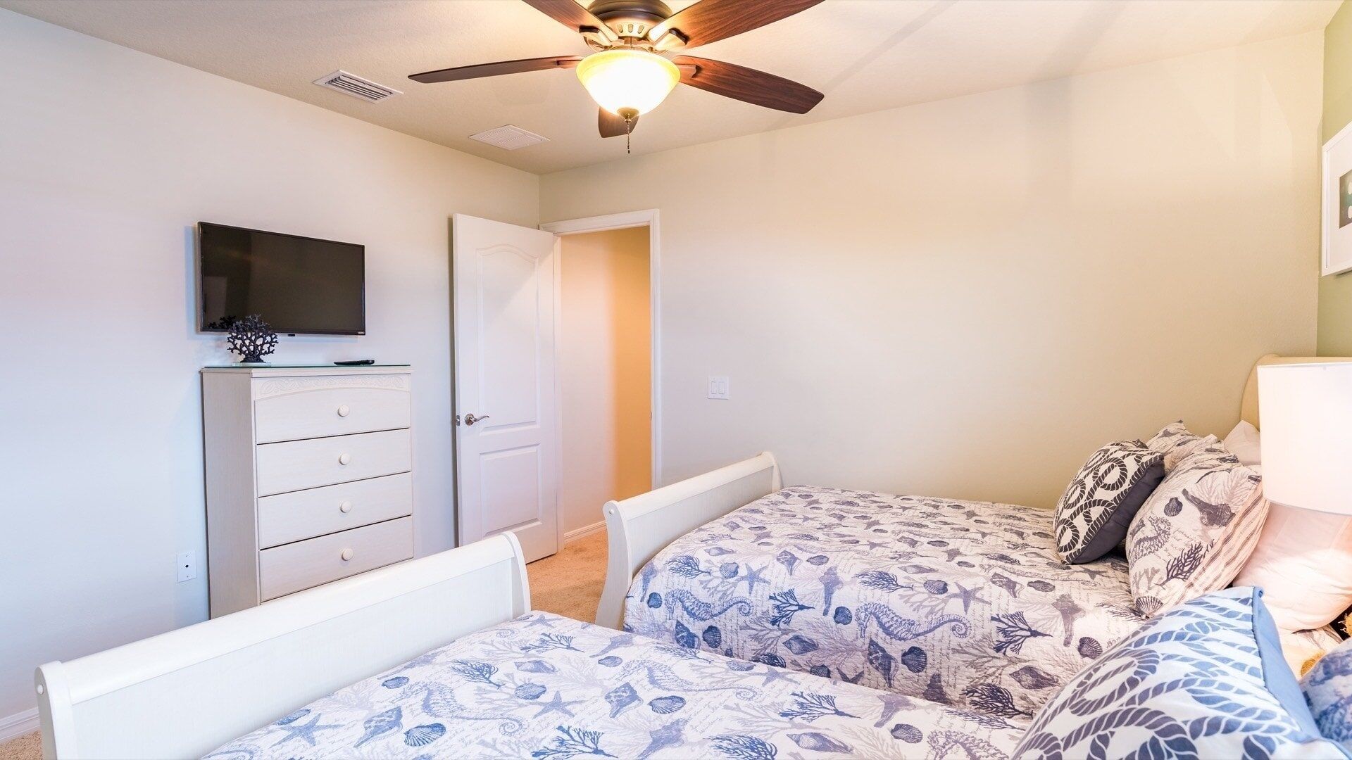 Een slaapkamer met twee bedden, een plafondventilator, een televisie op een dressoir, en een deur naar een andere kamer.