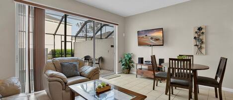 A spacious living room with a sliding glass door leading to an outdoor area, a comfortable sofa, a coffee table, a dining table with chairs, and a flat-screen TV mounted on the wall.