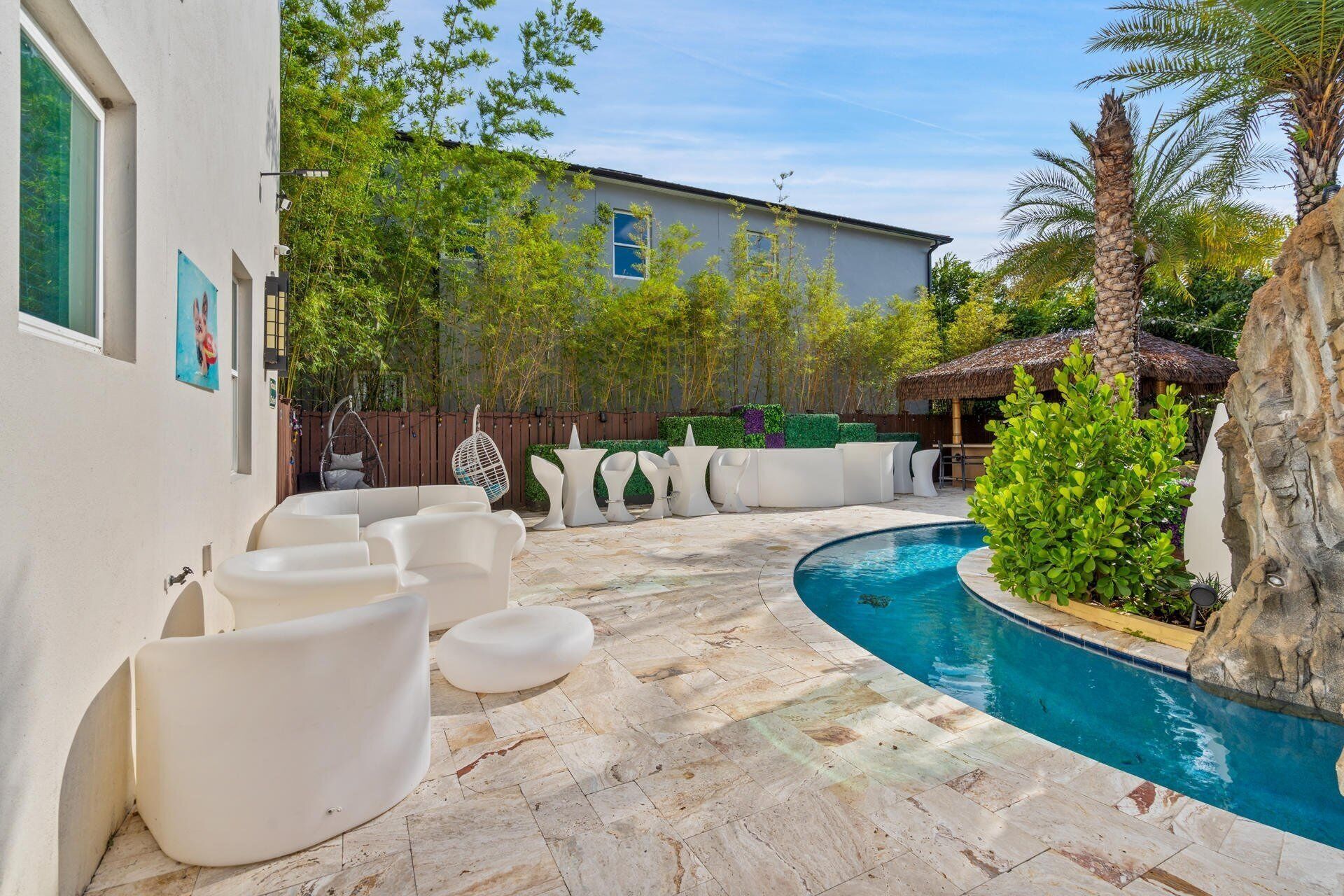 Zona de piscina con mobiliario exterior blanco, suelo de piedra y una piscina rodeada de vegetación y un edificio al fondo.