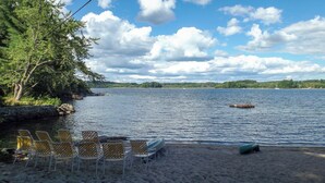 A beach with chairs and a small dock extending into the water, surrounded by trees and a distant shoreline. - Rare 4-Home Compound on Sebago Lake Peninsula With 360° Views (Raymond)