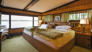 A large bed with a headboard, a bedside table with a lamp, a window with curtains, and a view of a lake. - Rare 4-Home Compound on Sebago Lake Peninsula With 360° Views (Raymond)