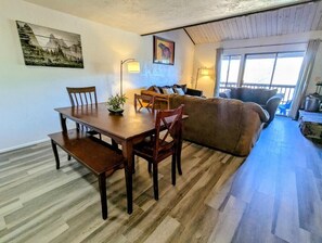 Ein gemütliches Zimmer mit einem Holzesstisch, Stühlen, einem Sofa und einer Sitzecke. An den Wänden hängen zwei Bilder und ein Fenster mit Vorhängen.