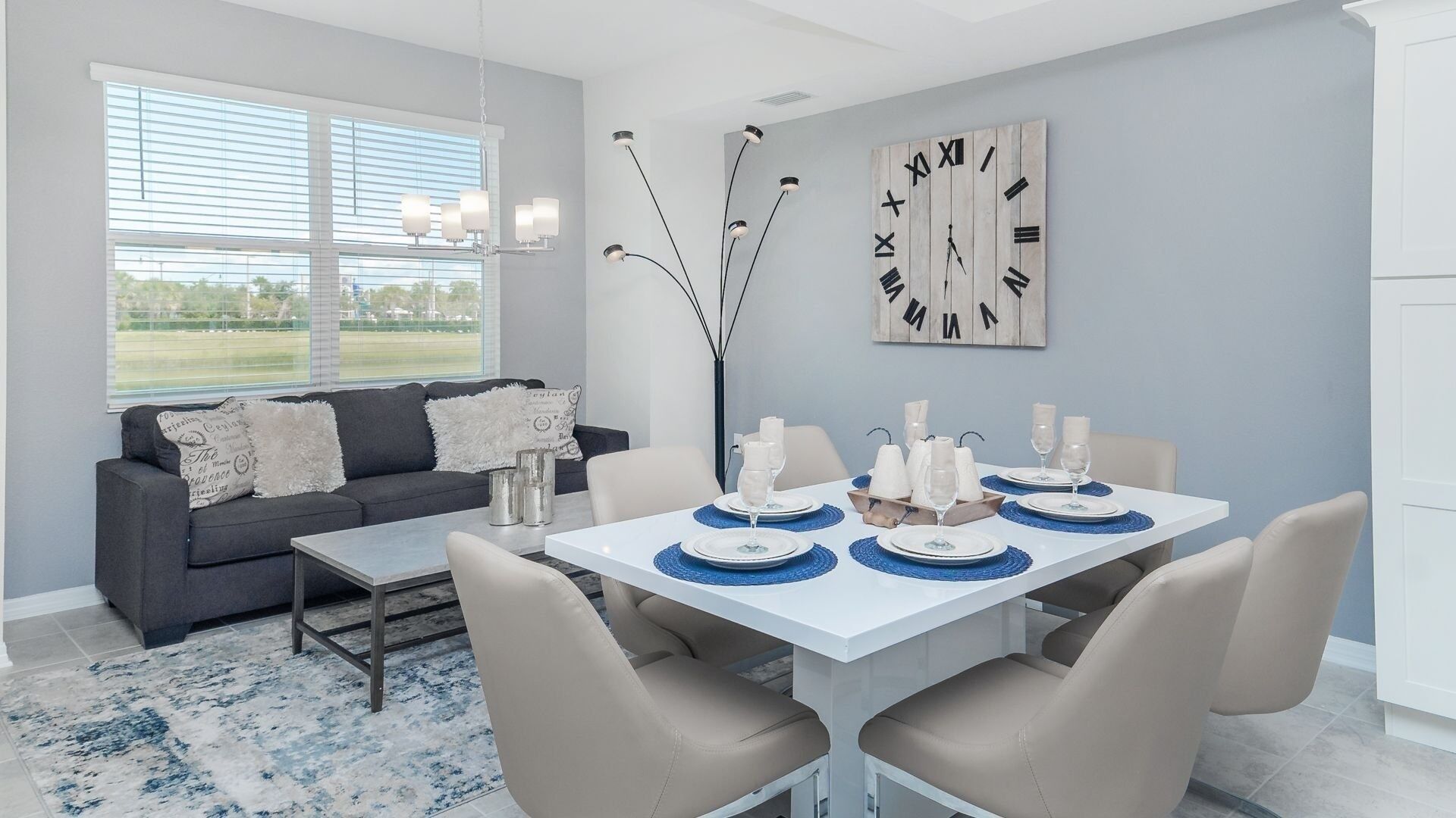 A modern dining area with a white table set for six, a grey sofa with decorative pillows, and a large wall clock.