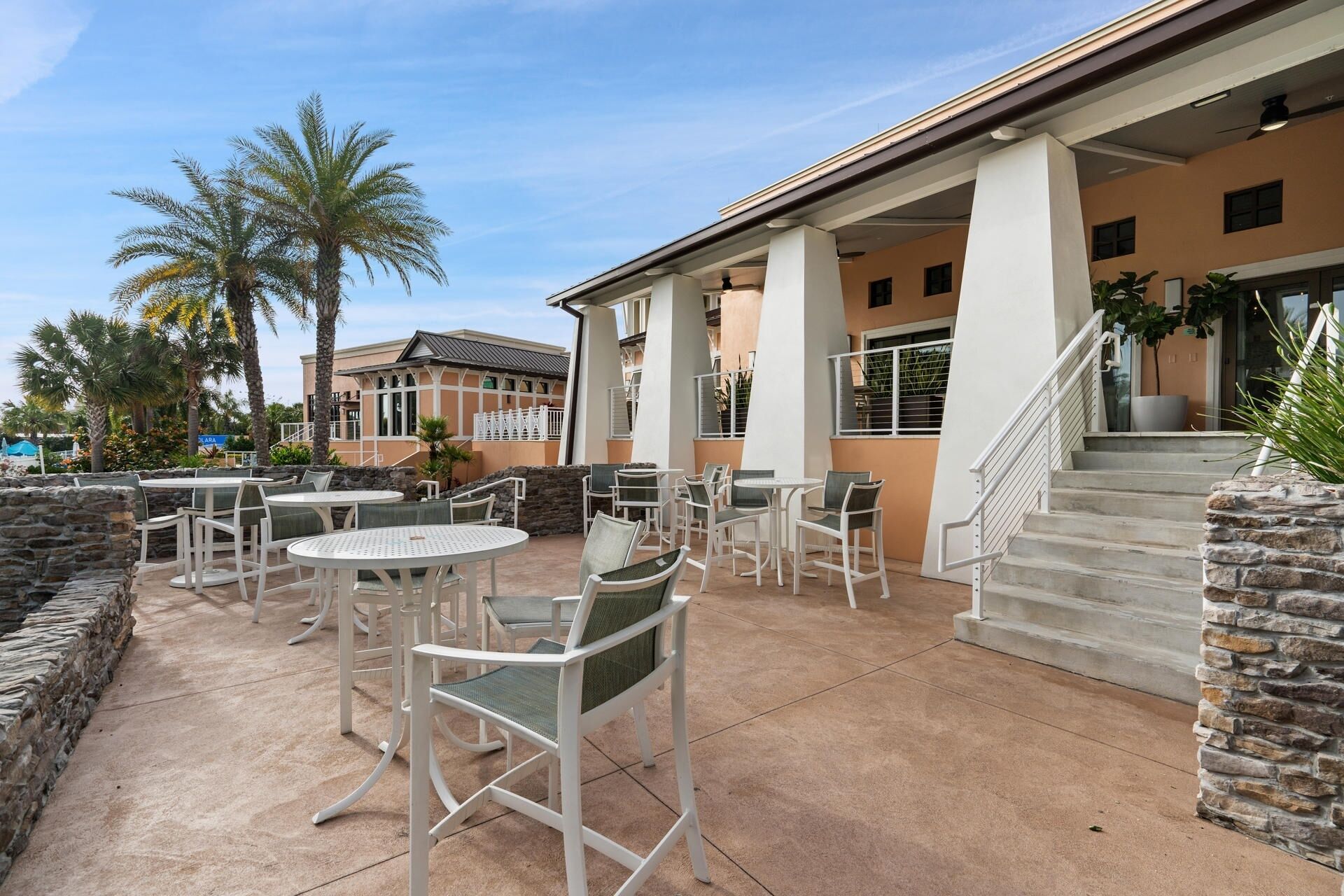 An outdoor patio area with round tables and chairs, stone walls, and palm trees.