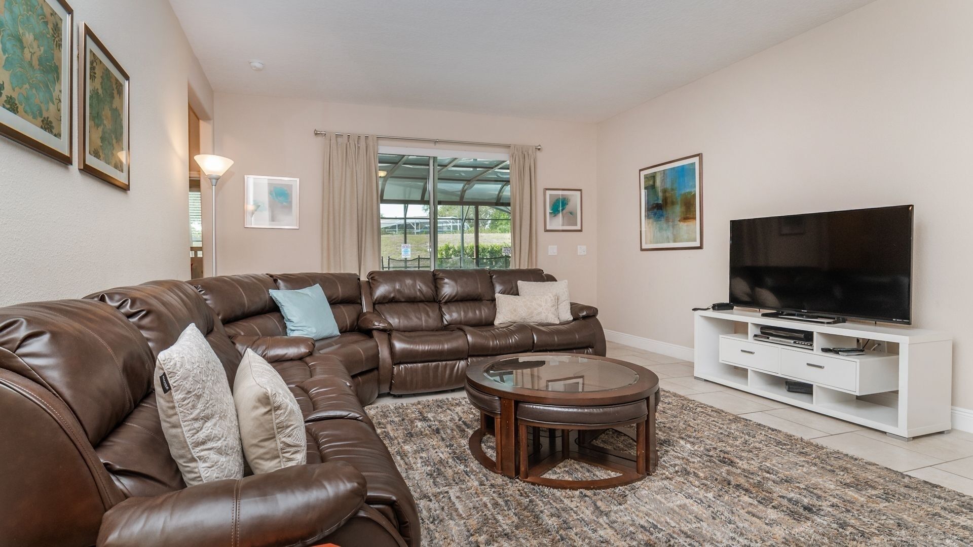 A living room with a brown leather sectional sofa, a glass coffee table, a flat-screen TV on a white entertainment unit, and artwork on the walls.