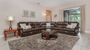 A living room with a brown leather sectional sofa, a round coffee table, a wooden side table, and a patterned rug. There are two framed artworks on the wall, a floor lamp, and a large window with curtains. - Solterra Resort 5 Bedroom Pool Home Pet Friendly 5 Home (Davenport)