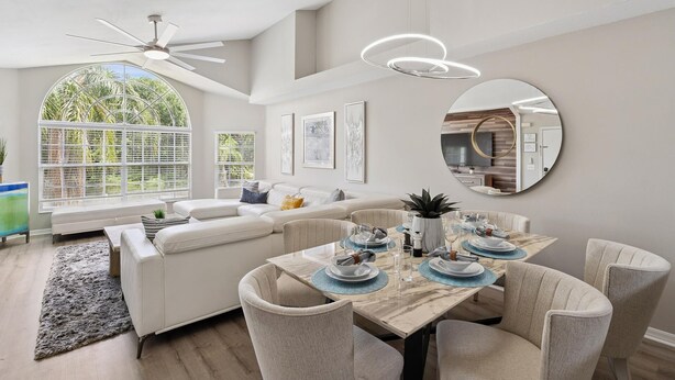 A modern living room with a dining area, featuring a large window, ceiling fan, and a round mirror. - Royal Palm Bay 3-Bedroom Vacation Condo with Amenity Access 3 Condo (Kissimmee)
